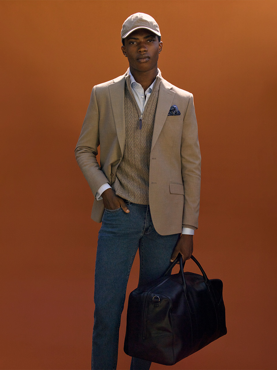 Male model wearing a camel blazer layered over a textured knit and shirt, styled with denim jeans and a tan cap, holding a black leather duffle bag against a burnt orange backdrop. Smart casual look from the Politix Autumn Winter 2025 Collection.