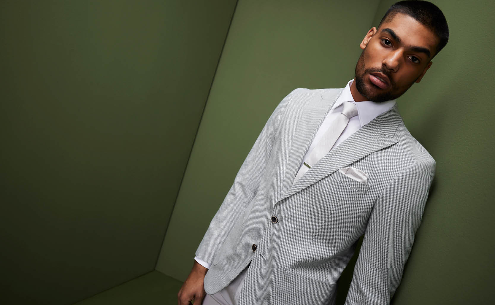 Male model wearing POLITIX light grey suit and white accessories standing in front of dark green backdrop