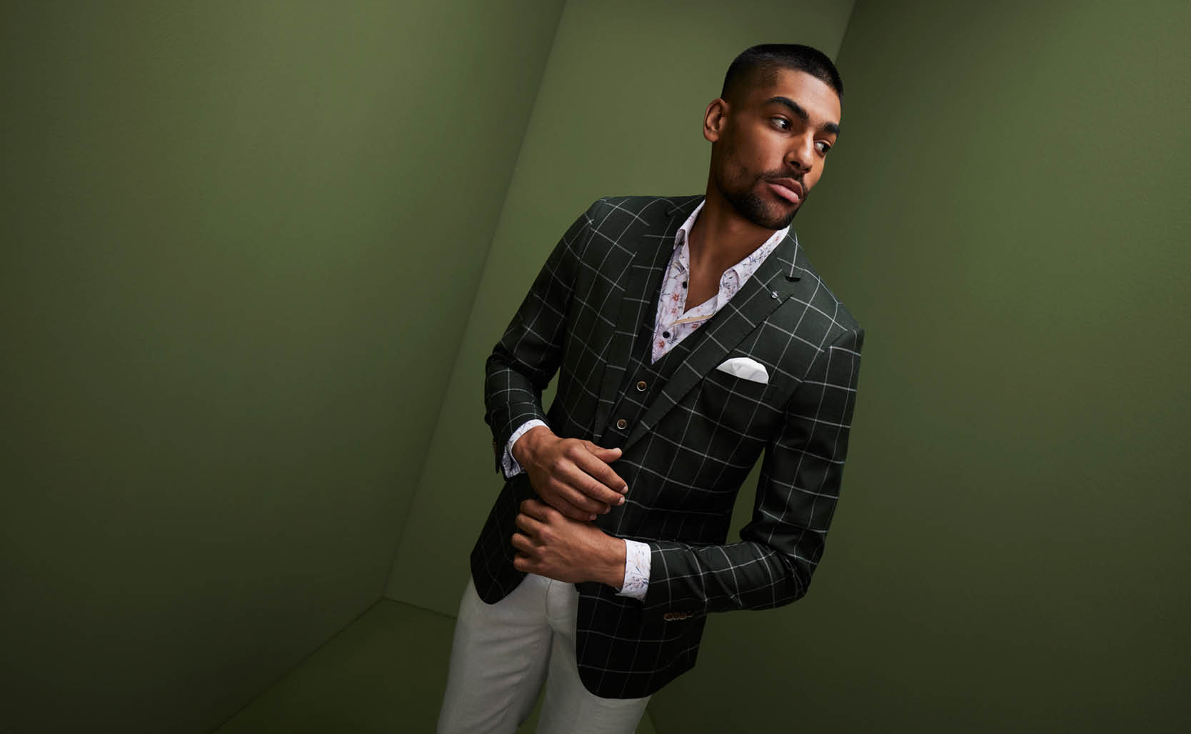 Model looking away in front of green wall wearing dark dress POLITIX suit jacket with white windowpane check, matching vest and floral button up shirt 