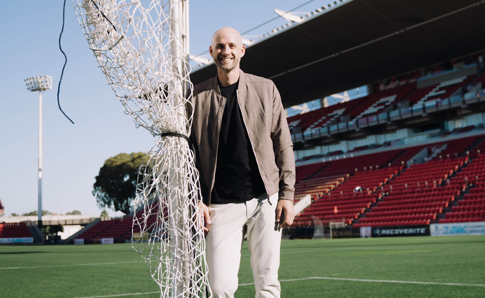 Jarrod Walsh featuring POLITIX Jarrod Walsh smiling standing next to a soccer post wearing beige bomber jacket, black shirt and white chinos
