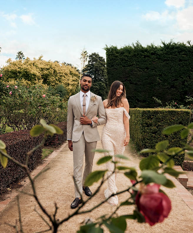 Male model wearing beige suit with female moderl wearing glittery weading dress walking through winery garden setting