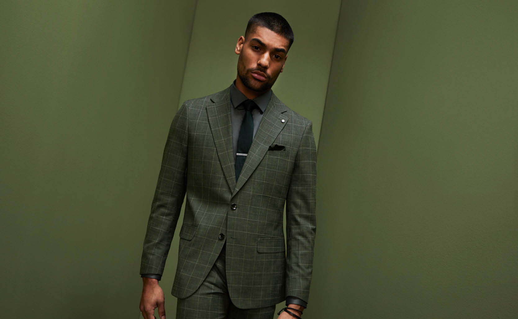Male model in POLITIX dark green windowpane suit with green tie and shirt standing in front of green wall