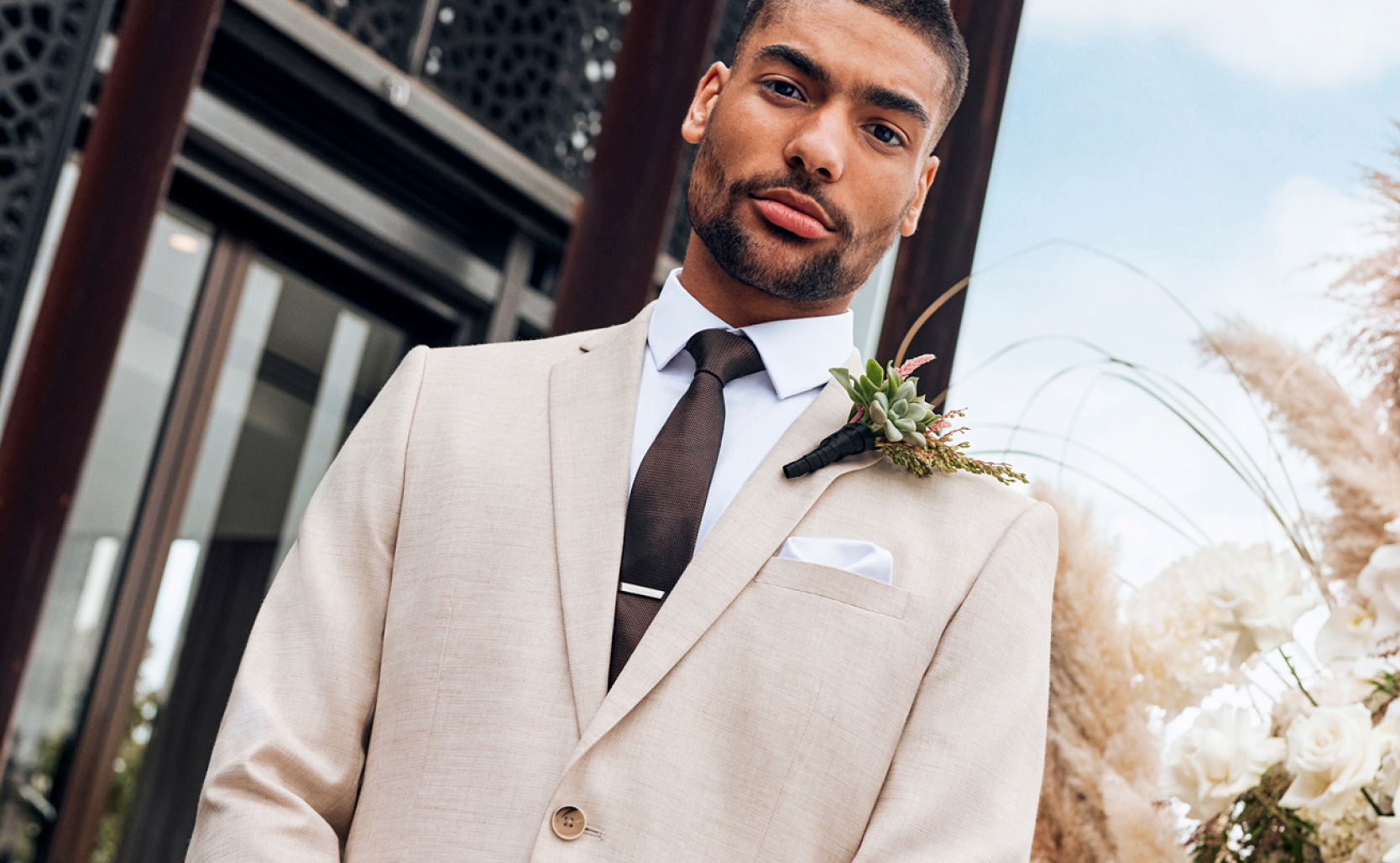 Image of model on hotel deck looking into camera wearing natural coloured suit with White button up shirt and burgundy tie
