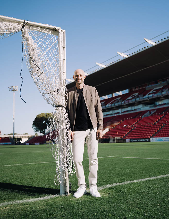Jarrod Walsh featuring POLITIX Jarrod Walsh smiling standing next to a soccer post wearing beige bomber jacket, black shirt and white chinos
